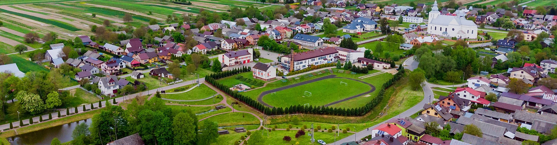 Zielone boisko sportowe otoczone drzewami, domy jednorodzinne o różnorodnych dachach, biała duża budowla z wieżą, pola uprawne w tle.