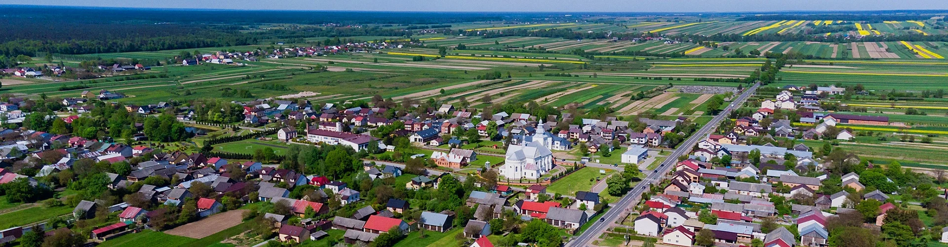 Widok z lotu ptaka na wieś z licznymi domami, białym kościołem w centrum, drogą i zielonymi polami oraz żółtymi pasami kwitnących roślin w tle.