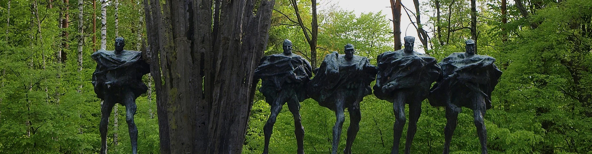 Pięć ciemnych, abstrakcyjnych postaci z rozłożystymi ramionami na kamiennej podstawie, w tle gęsty zielony las i wysoki, rozgałęziony pień drzewa.