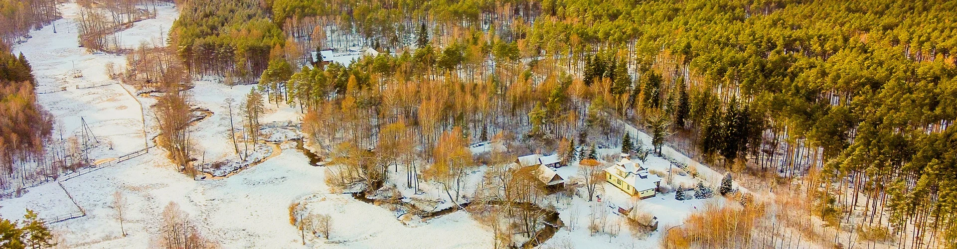 Zima w lesie z domem i budynkami pokrytymi śniegiem, wokół drzewa iglaste i liściaste, częściowo ośnieżona polana, strumień.