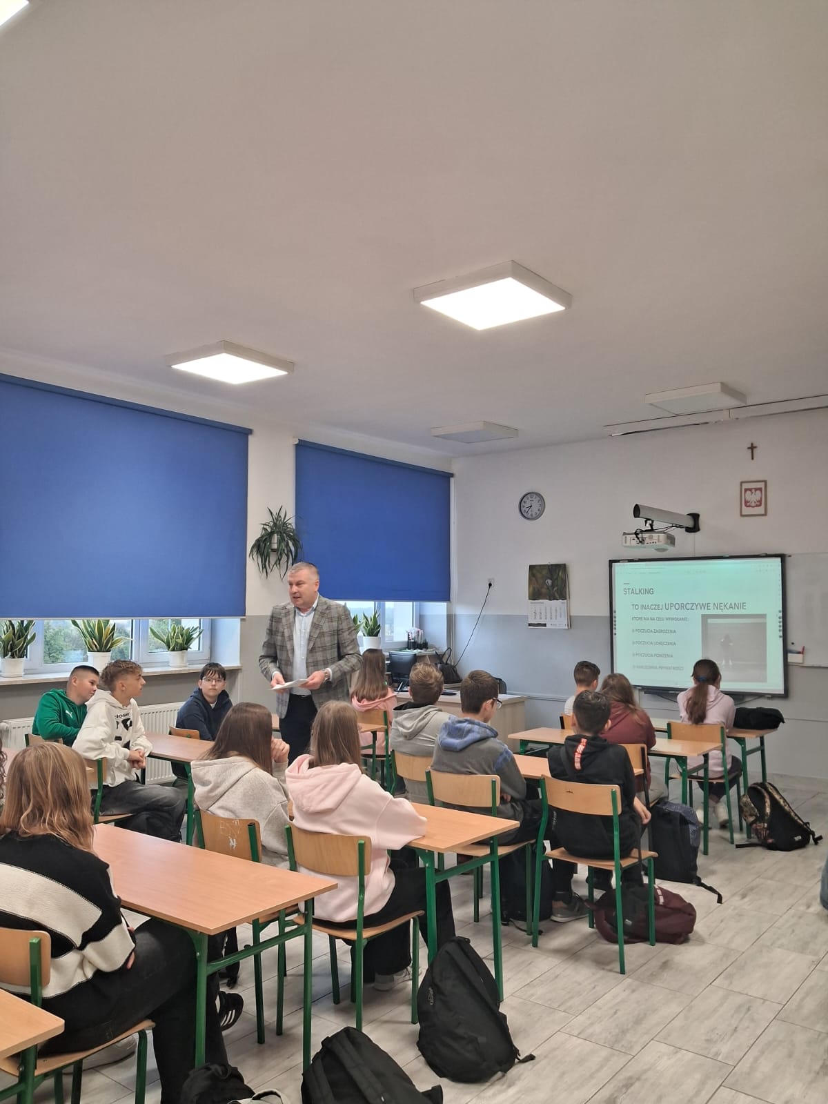 Klasa szkolna z uczniami siedzącymi przy drewnianych ławkach i nauczycielem w szarej marynarce, niebieskie rolety na oknach, tablica multimedialna z wyświetlonym tekstem.