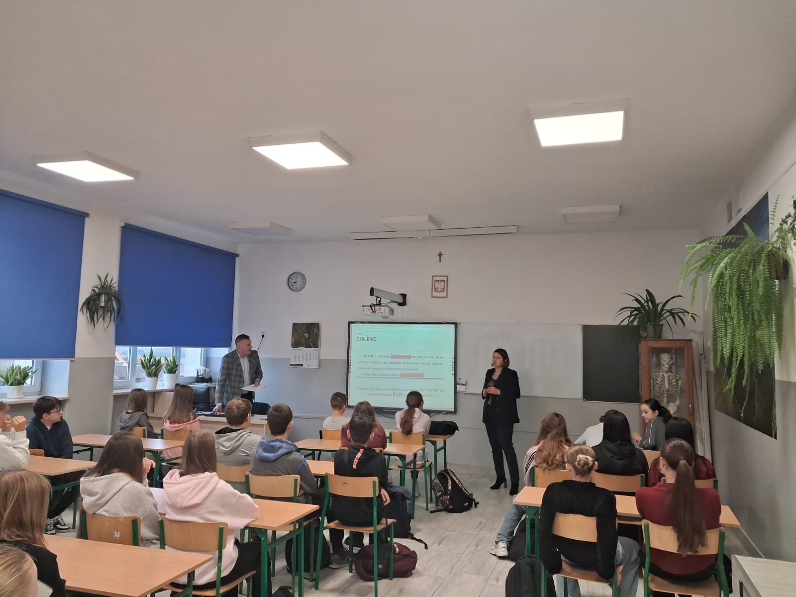 Sala lekcyjna z uczniami siedzącymi przy ławkach, nauczycielami przy tablicy, niebieskie rolety na oknach, rośliny na parapetach i ścianach, na ścianie szkielet w gablocie.