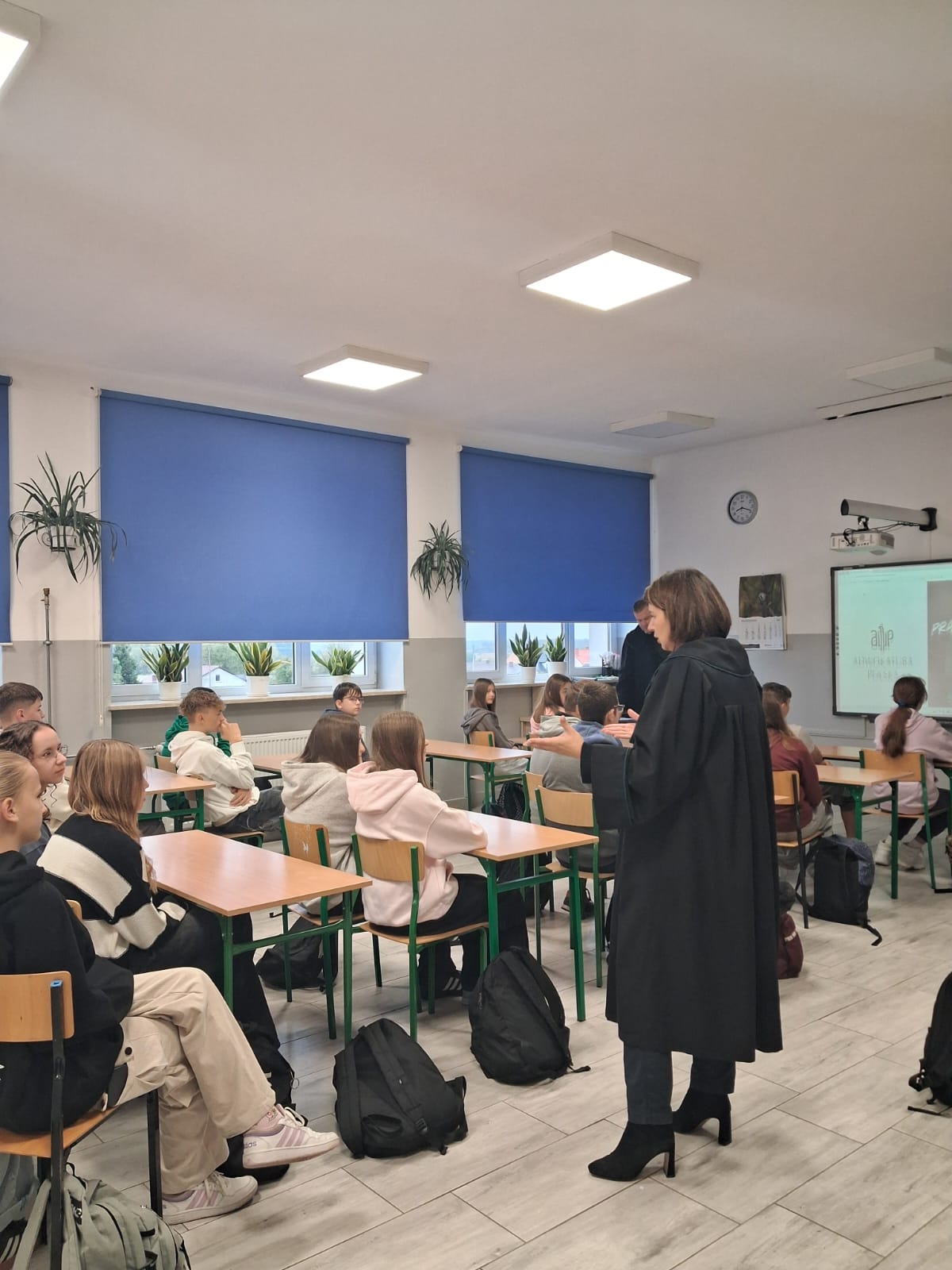Klasa szkolna z uczniami siedzącymi przy drewnianych ławkach z zielonymi metalowymi nogami, niebieskie rolety na oknach, nauczycielka w czarnym płaszczu stoi obok.
