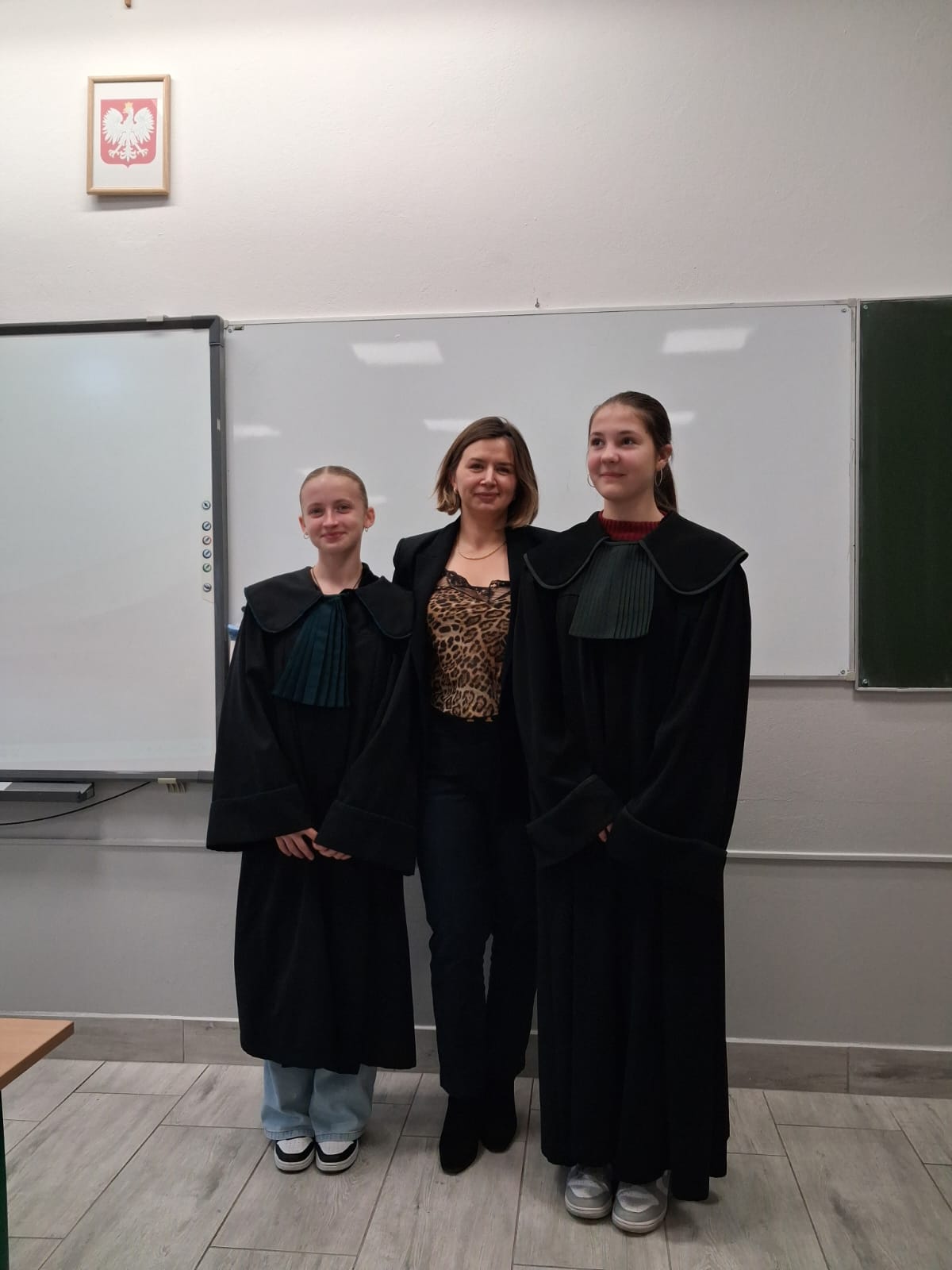 Trzy kobiety stoją na tle białej tablicy, dwie w długich czarnych togach z plisowanymi kołnierzami, jedna w czarnym garniturze i panterkowej bluzce.