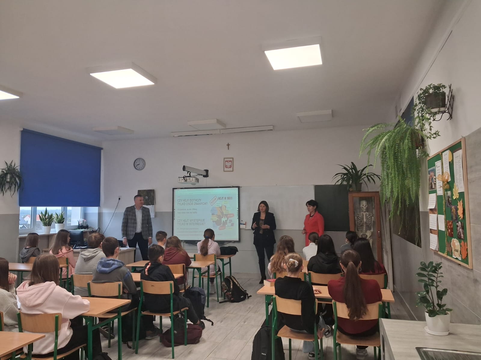 Klasa szkolna z uczniami siedzącymi przy drewnianych ławkach, trzy osoby stojące przy tablicy, zielone rośliny doniczkowe, niebieska roleta, ekran z projekcją.