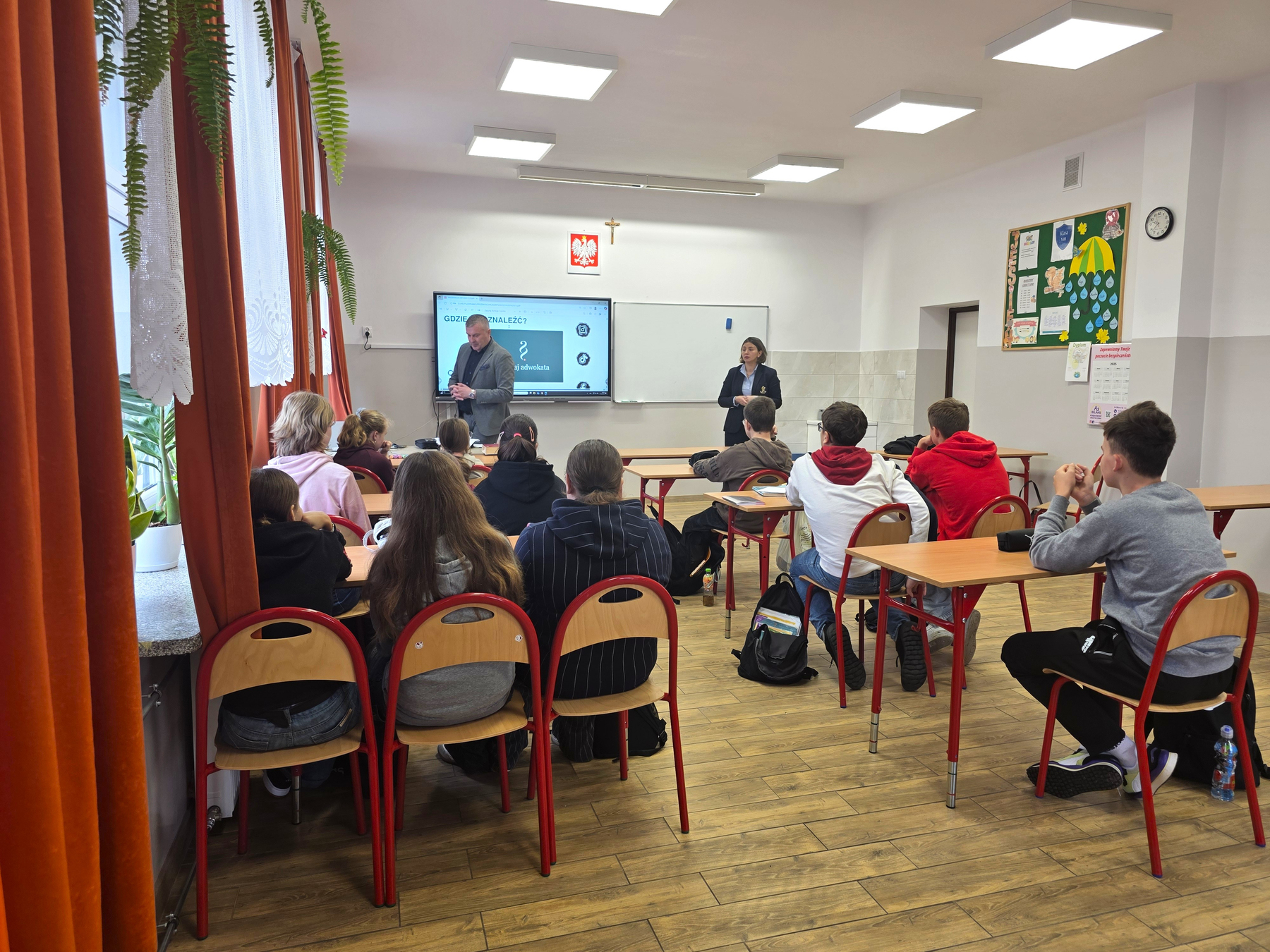Klasa szkolna z uczniami siedzącymi na krzesłach z czerwonymi nogami, dwie osoby prowadzące lekcję, białe ściany, drewniana podłoga, zielona tablica korkowa, czerwone zasłony przy oknie.