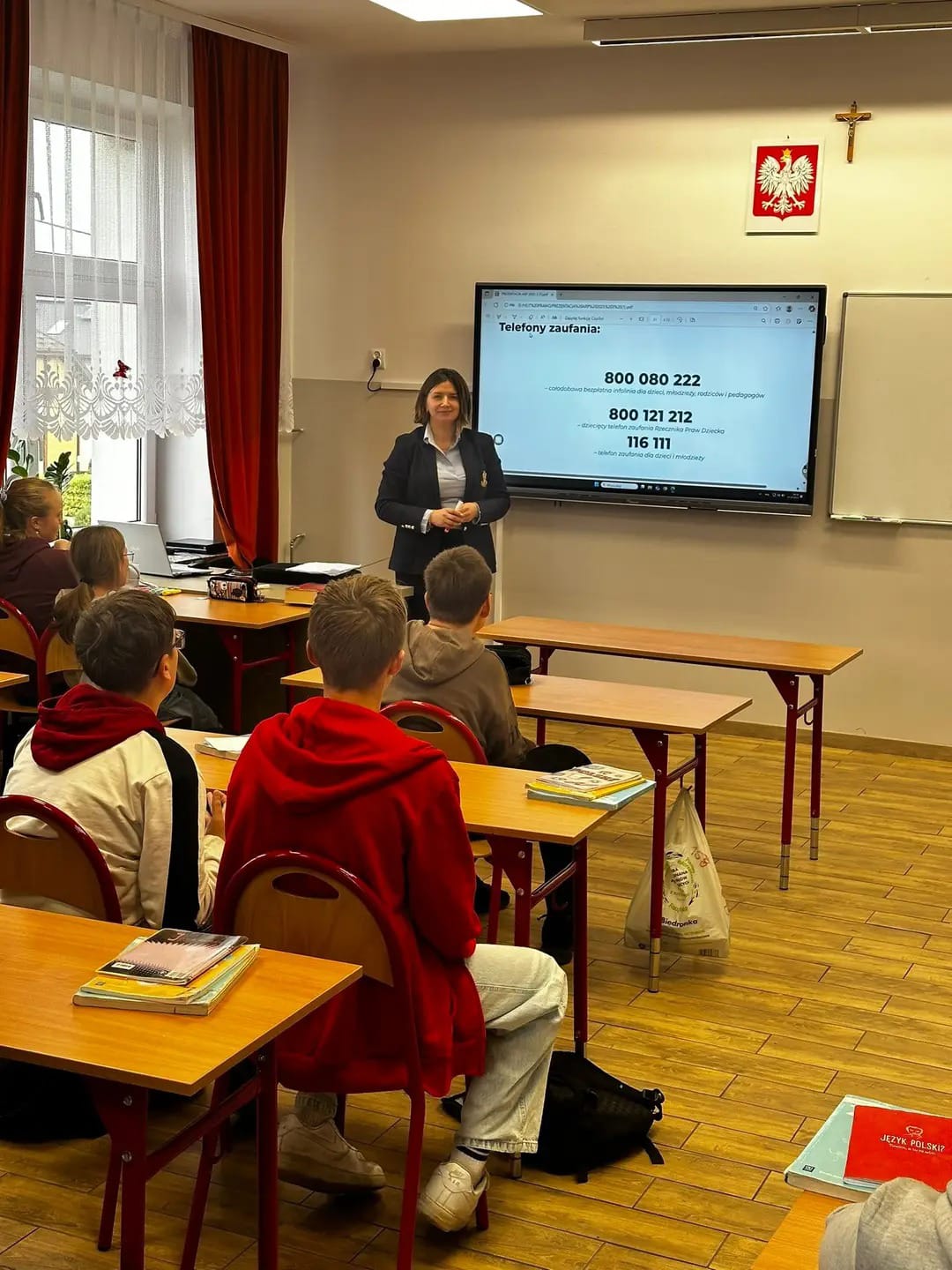Sala lekcyjna z uczniami siedzącymi przy drewnianych ławkach, nauczycielka przed ekranem z wyświetlonymi numerami telefonów zaufania, na ścianie godło Polski i krzyż.