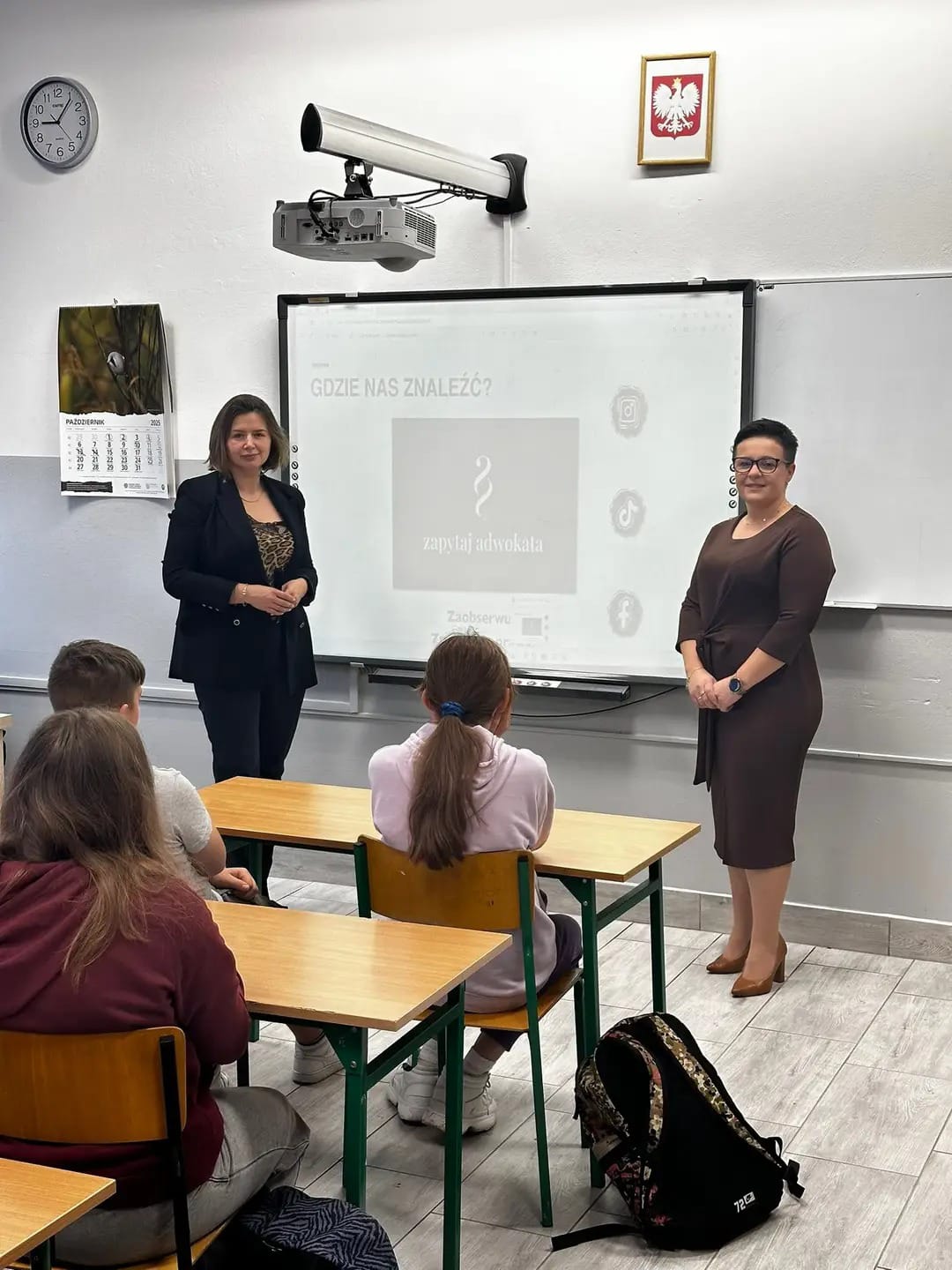 Klasa szkolna z uczniami siedzącymi przy drewnianych ławkach, dwie kobiety stojące przy tablicy interaktywnej, ściana z kalendarzem, zegarem i godłem Polski.