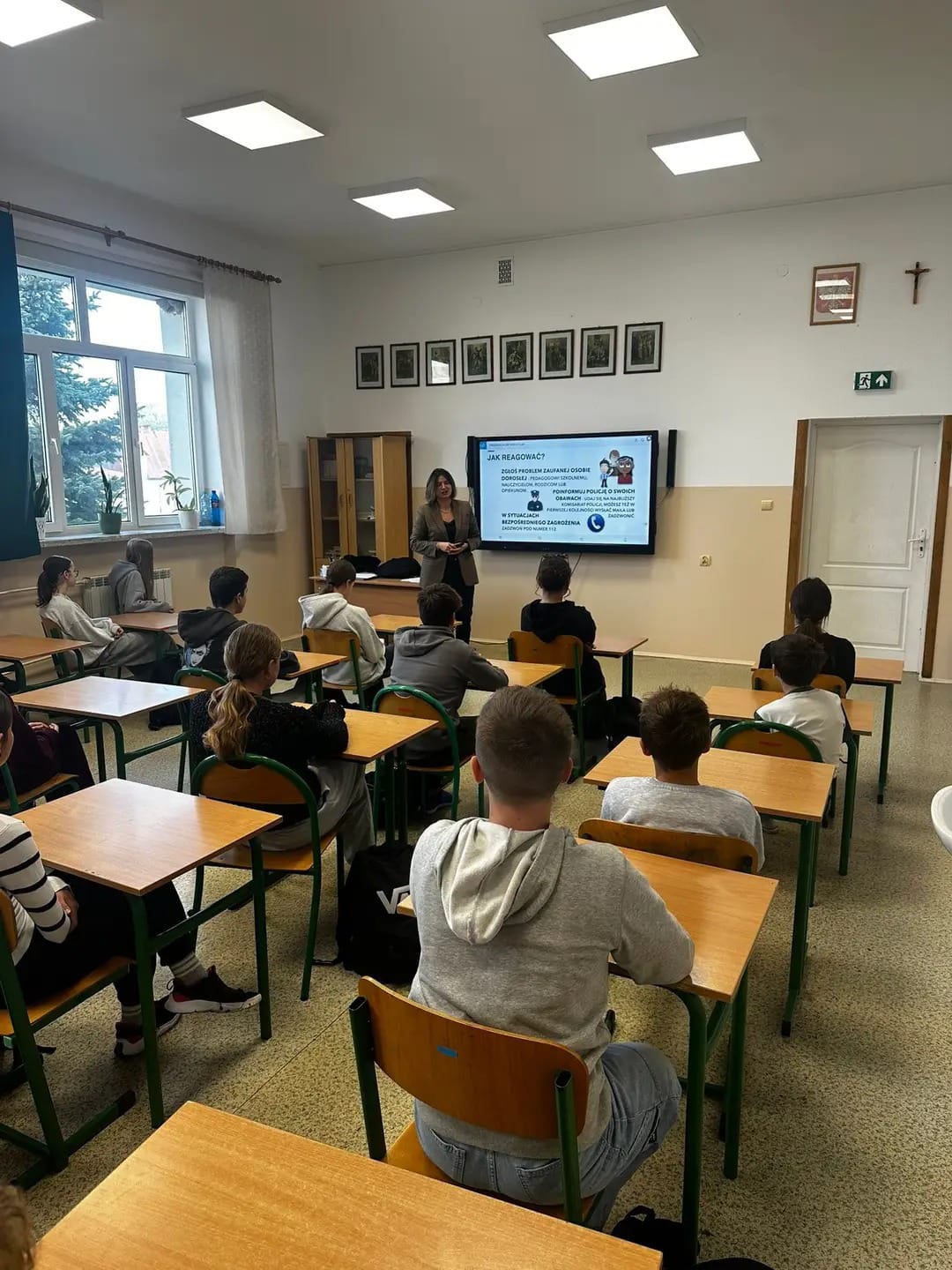 Uczniowie siedzący przy drewnianych ławkach w klasie, nauczycielka stoi przy tablicy multimedialnej z prezentacją, jasne oświetlenie, okno z roślinami, białe ściany.