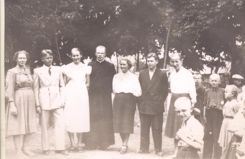 Grupa ludzi w strojach z dawnych lat na tle drzew, wśród nich mężczyzna w sutannie, kobiety w sukienkach i mężczyźni w garniturach, część dzieci z boku.