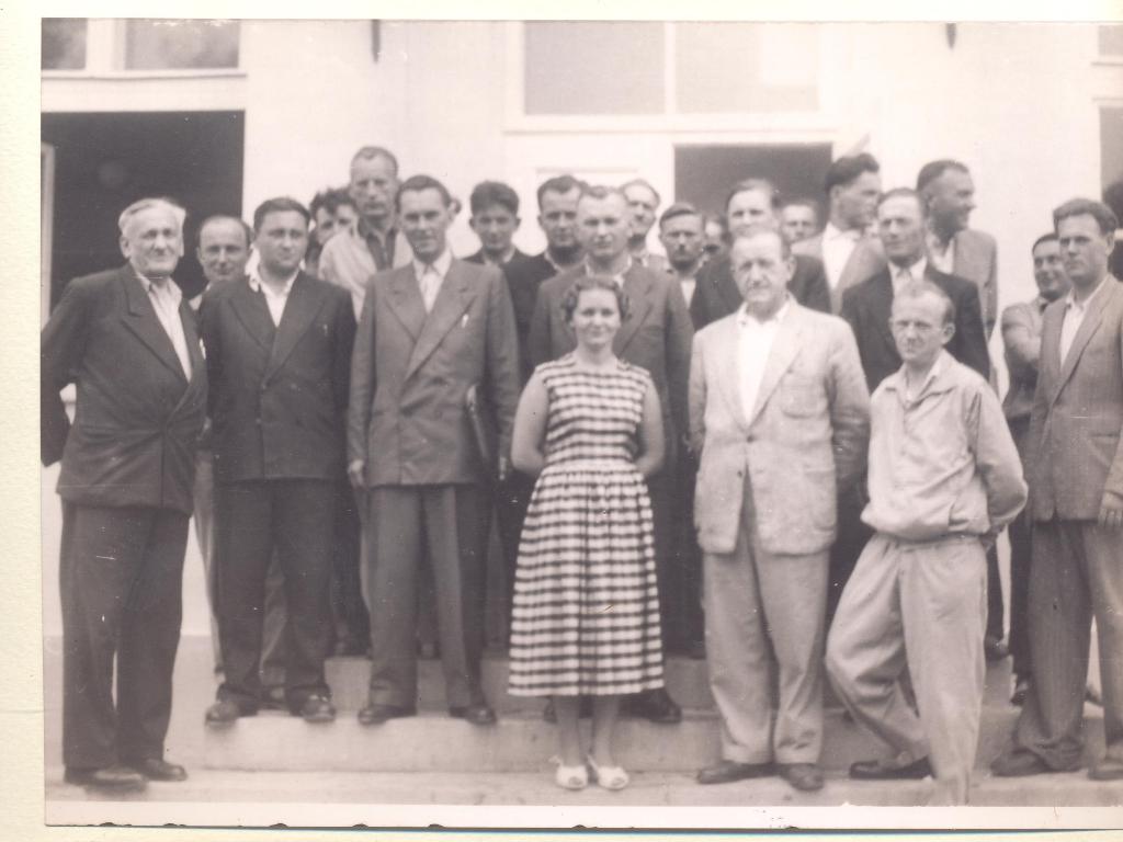 Grupa mężczyzn i kobieta na schodach przed jasnym budynkiem, mężczyźni w garniturach lub ubraniach roboczych, kobieta w sukience w poziome pasy.