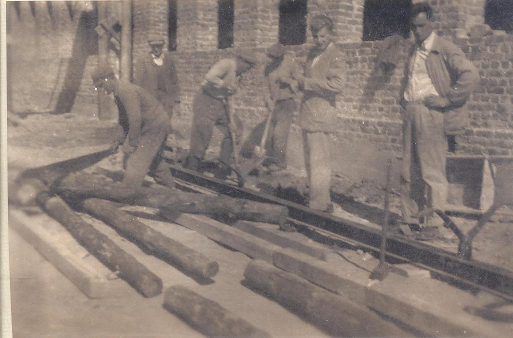 Grupa mężczyzn przy murze z cegieł, układająca lub przesuwająca długie drewniane kłody na ziemi, ubrani w robocze stroje, obraz w sepii.