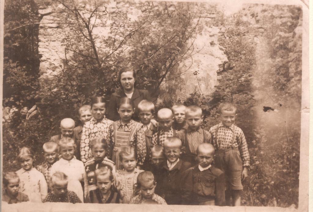 Grupa dzieci w różnorodnych ubraniach, stojących przed ścianą z roślinnością, w tle kobieta w ciemnej marynarce, zdjęcie w tonacji sepii.