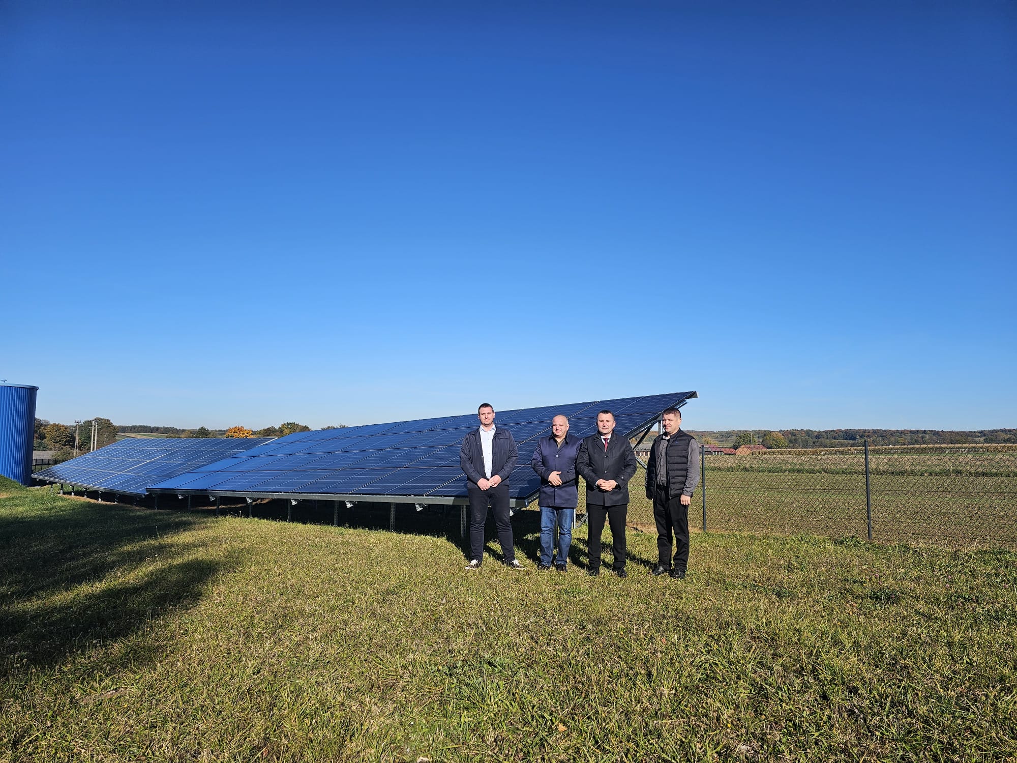 Czterech mężczyzn stoi na zielonej trawie obok pochylonych paneli słonecznych, za nimi łańcuchowy płot, w tle pola i las pod bezchmurnym niebem.