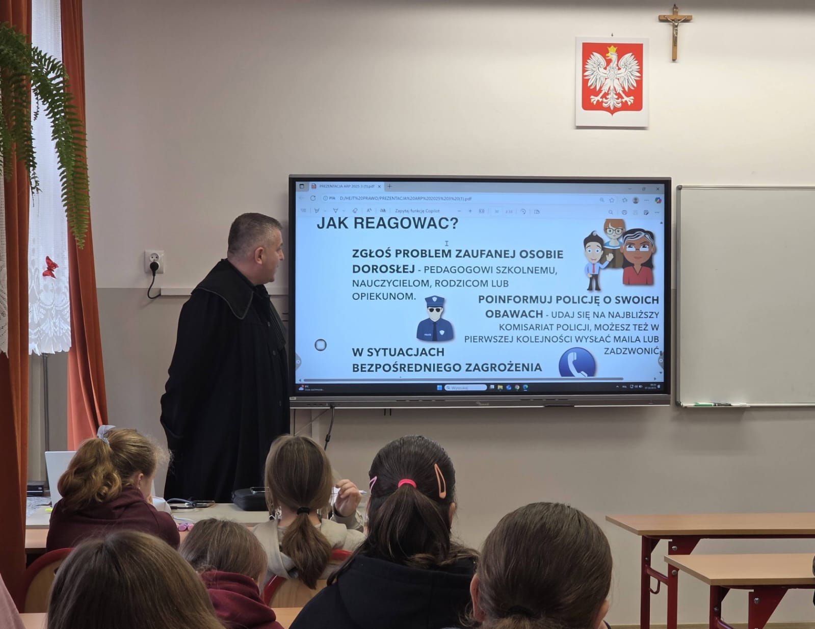 Mężczyzna w czarnym ubraniu stoi przy ekranie z tekstem w języku polskim, siedzące dzieci oglądają prezentację w klasie, na ścianie godło Polski i krzyż, zasłony pomarańczowe i roślina.