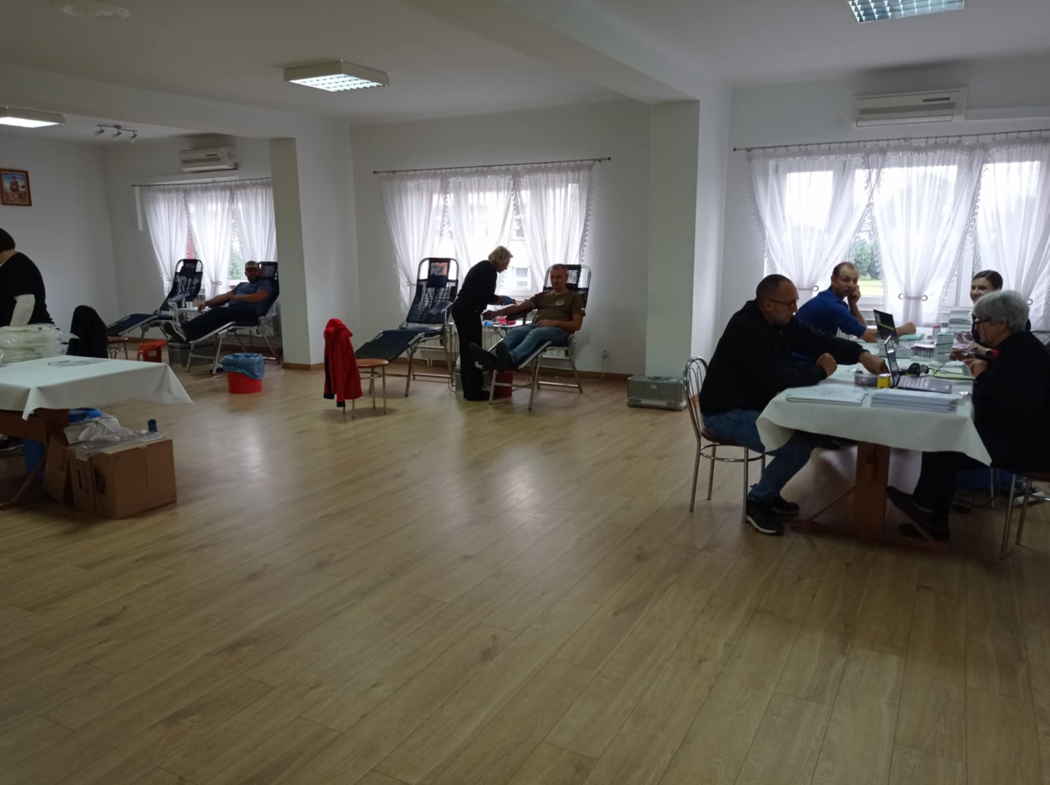 Wnętrze jasnego pomieszczenia z drewnianą podłogą, leżakami przy oknach z białymi firankami, ludźmi siedzącymi przy stołach i na leżakach, klimatyzatory na ścianach.