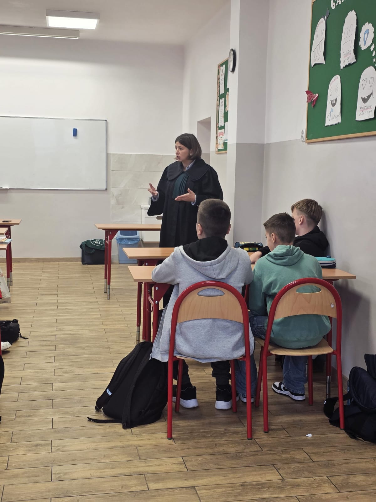 Klasowa sala z jasnymi ścianami, trzema siedzącymi chłopcami w bluzach i stojącą kobietą ubrana w ciemną szatę, przy drewnianych biurkach i krzesłach z czerwonymi ramami.