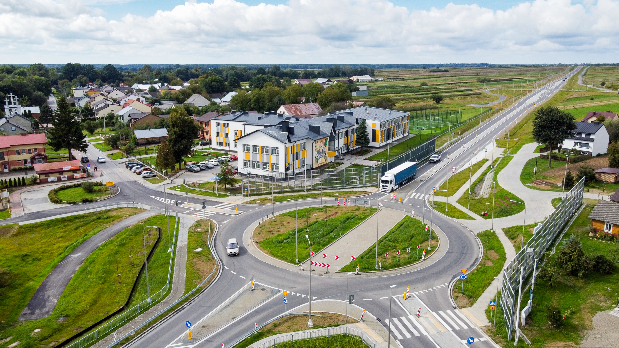 Rondo z dwoma pojazdami na drodze, budynek z muralem, zabudowa mieszkalna, zieleń, pola uprawne i niebieskie niebo z chmurami.