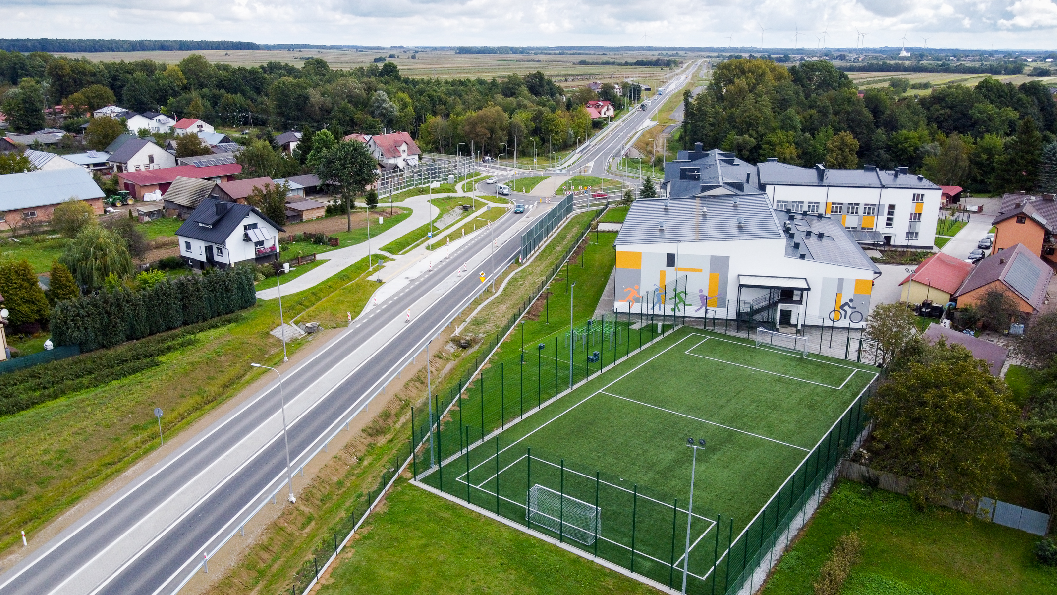 Boisko piłkarskie z zieloną sztuczną trawą, ogrodzone siatką, obok droga z kilkoma samochodami, budynki mieszkalne i szkoła z kolorowymi grafikami na ścianie, drzewa w tle.