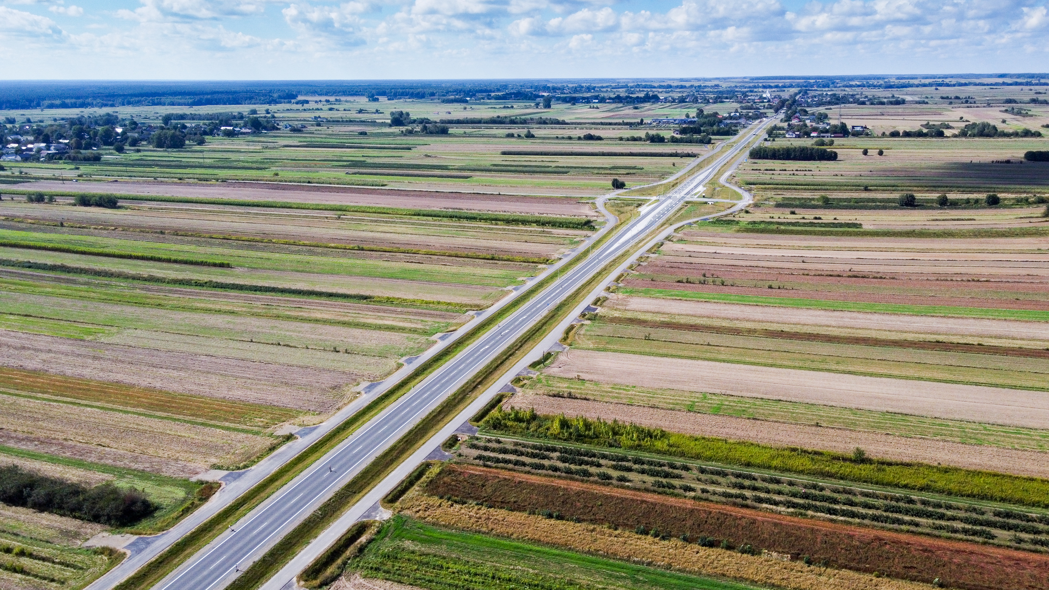 Droga przecinająca pola uprawne o różnych odcieniach zieleni, brązu i żółci, na horyzoncie zabudowania i las pod niebieskim niebem z chmurami.