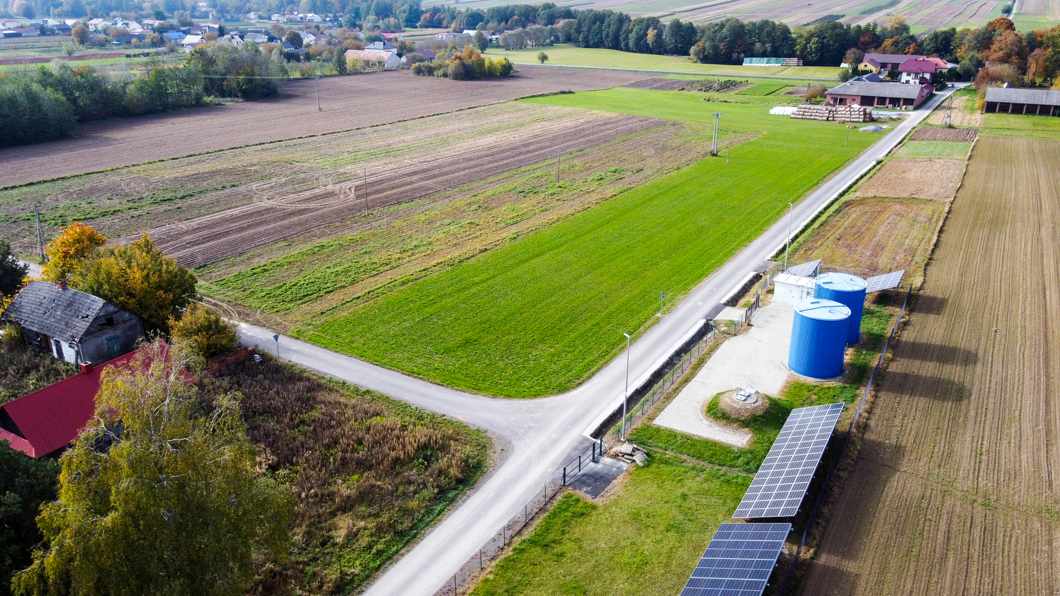 Zielone i brązowe pola uprawne, droga przecinająca teren, dwa niebieskie zbiorniki, panele słoneczne, zabudowania i drzewa o jesiennych barwach.