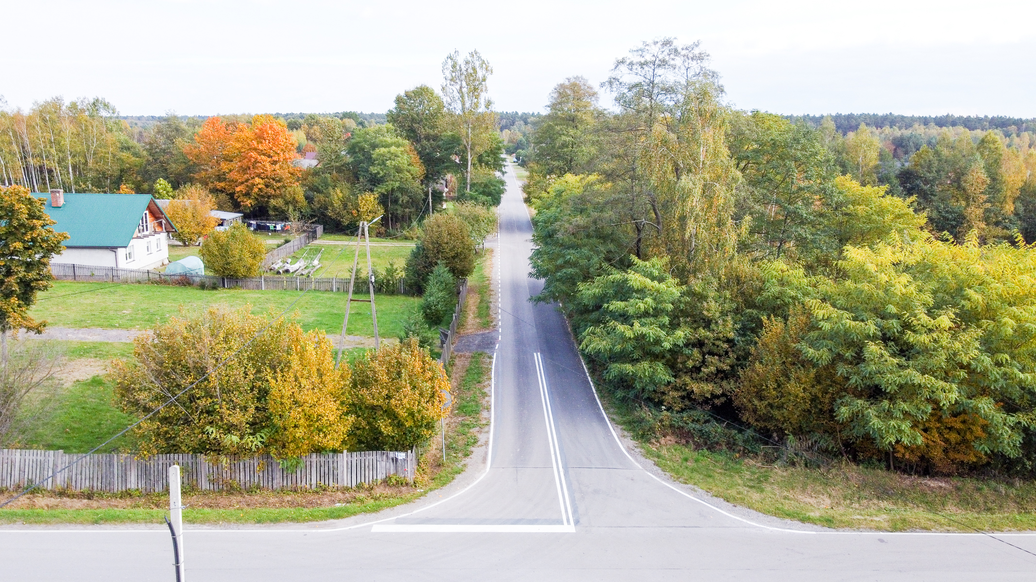 Droga asfaltowa z białym oznaczeniem, po lewej dom z zielonym dachem, ogrodzony płotem, wokół drzewa o jesiennych barwach zieleni, żółci i pomarańczu.