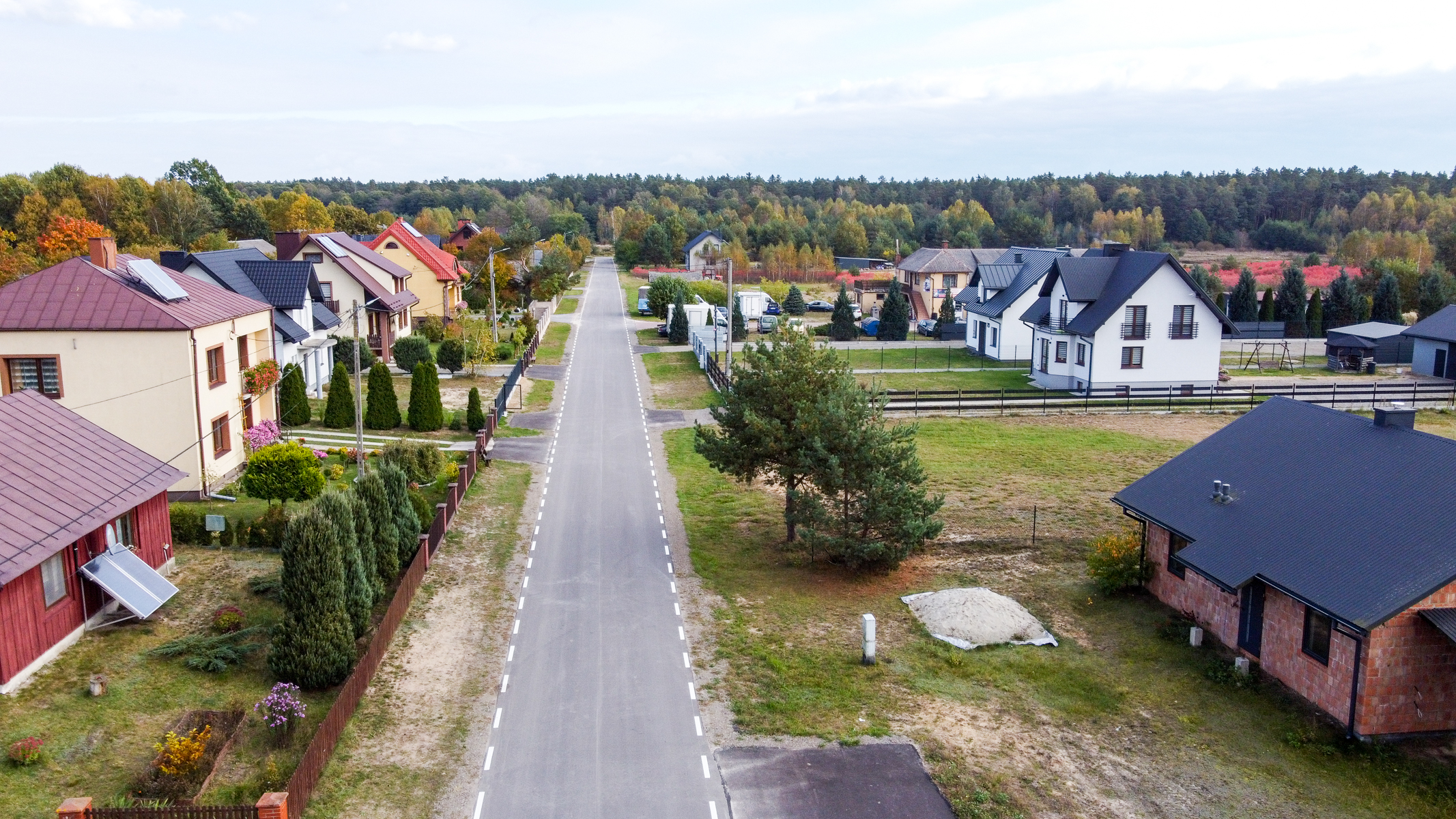 Widok z lotu ptaka na prostą, asfaltową drogę z domami po obu stronach, zielone trawniki, ogrodzenia oraz drzewa na tle lasu pod niebieskim niebem.