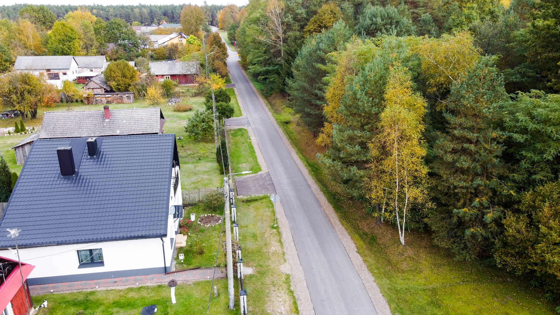 Droga asfaltowa wzdłuż domu z ciemnym dachem, po prawej stronie las z drzewami o zielonych i żółtych liściach, po lewej zabudowania i trawniki.