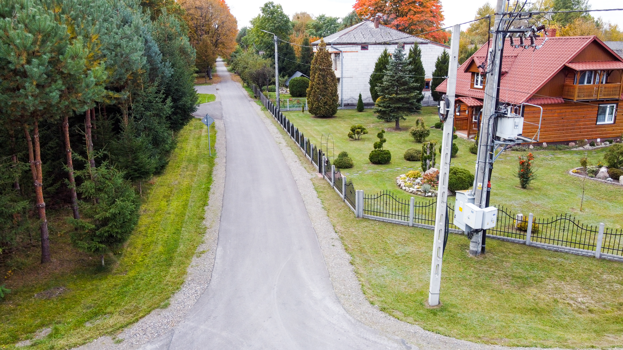 Asfaltowa droga z trawnikiem, po lewej gęste drzewa iglaste, po prawej domy z ogrodami, słup energetyczny z urządzeniami, jesienne drzewa w tle.