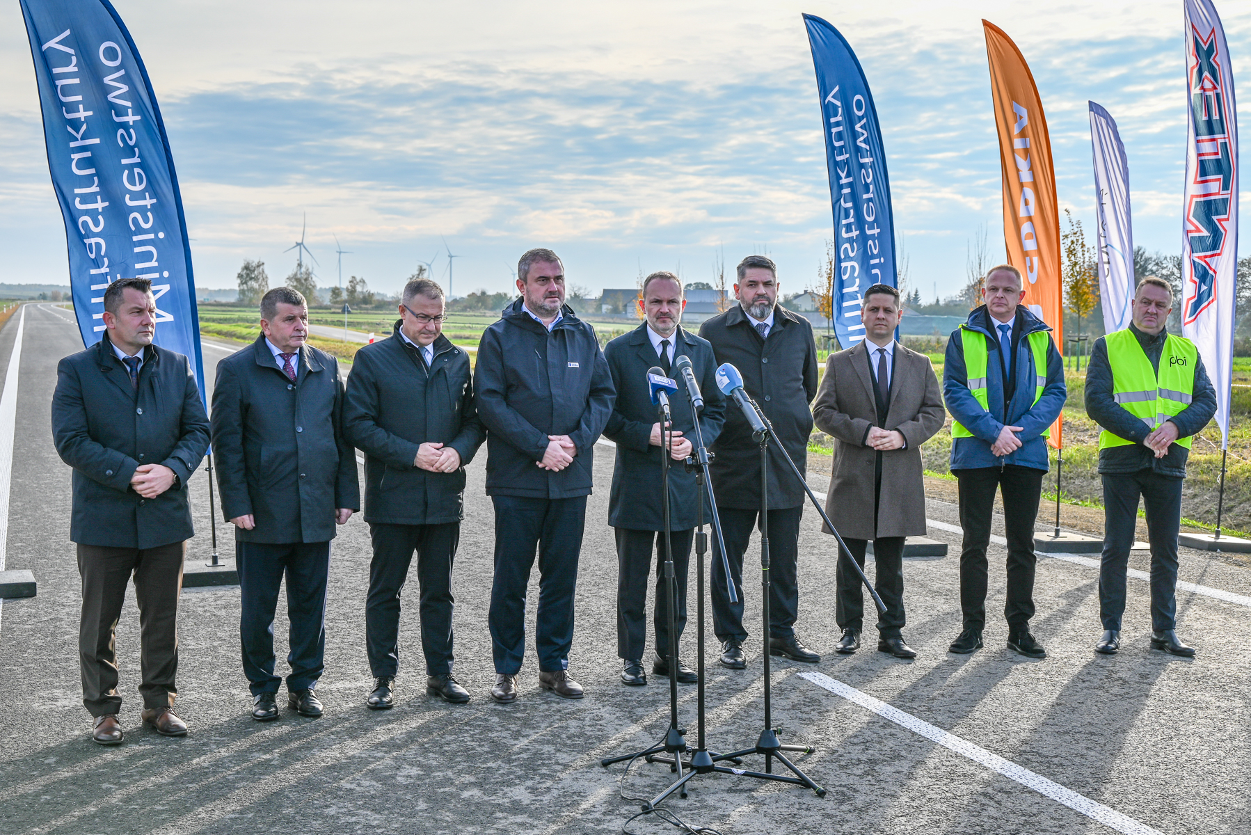 Dziesięciu mężczyzn w garniturach i kurtkach stoi na nowej drodze z mikrofonami, za nimi kolorowe flagi i pola z wiatrakami na horyzoncie.