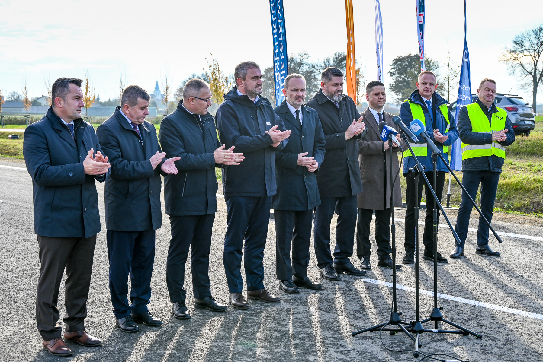 Dziesięciu mężczyzn w garniturach i kurtkach stoi na drodze, ośmiu z nich klaszcze, dwóch ma żółte kamizelki, na tle zielonych pól i flag.