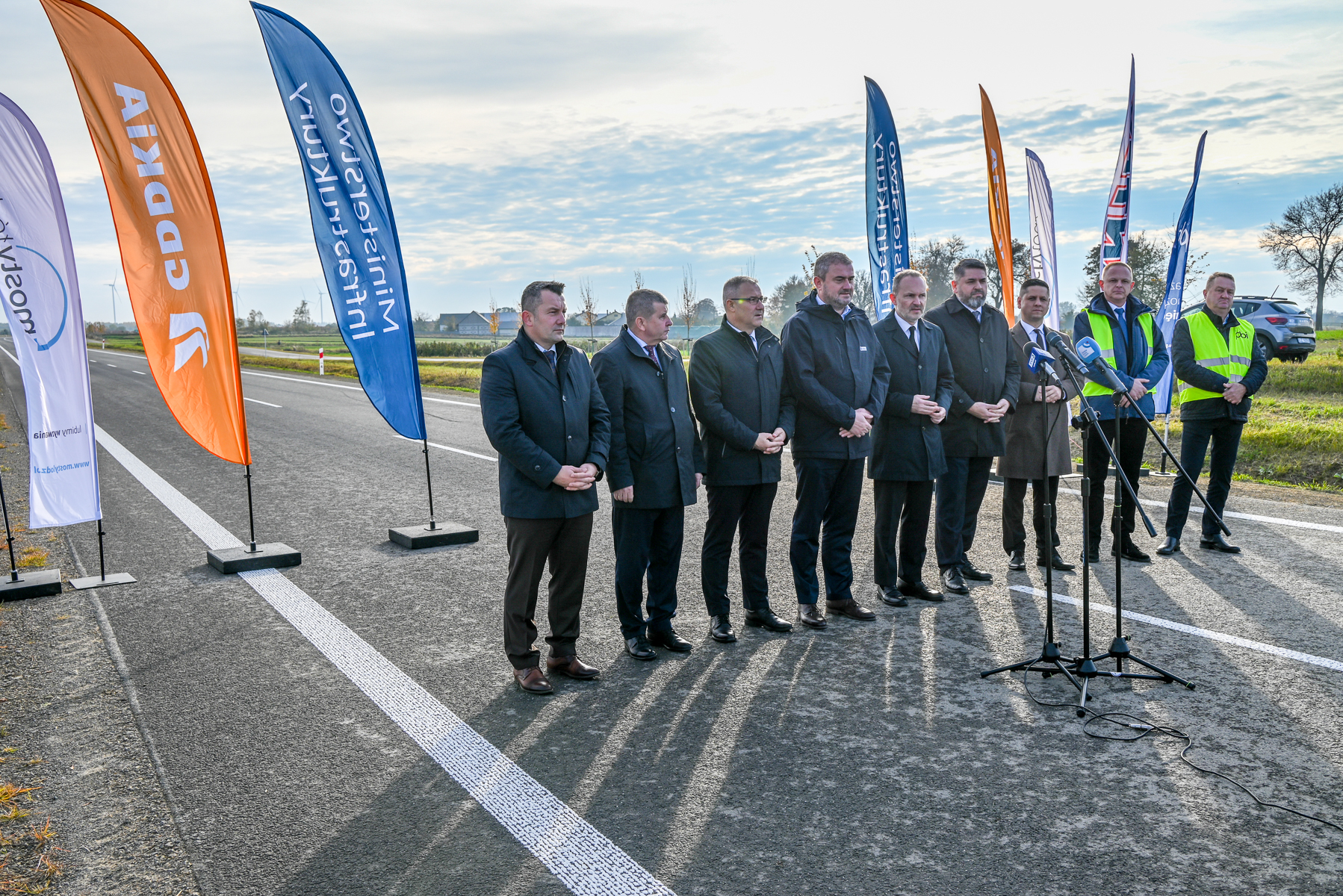 Grupa mężczyzn w ciemnych płaszczach i kamizelkach odblaskowych stoi na asfalcie drogi obok kolorowych flag i mikrofonów, w tle pola i częściowo zachmurzone niebo.