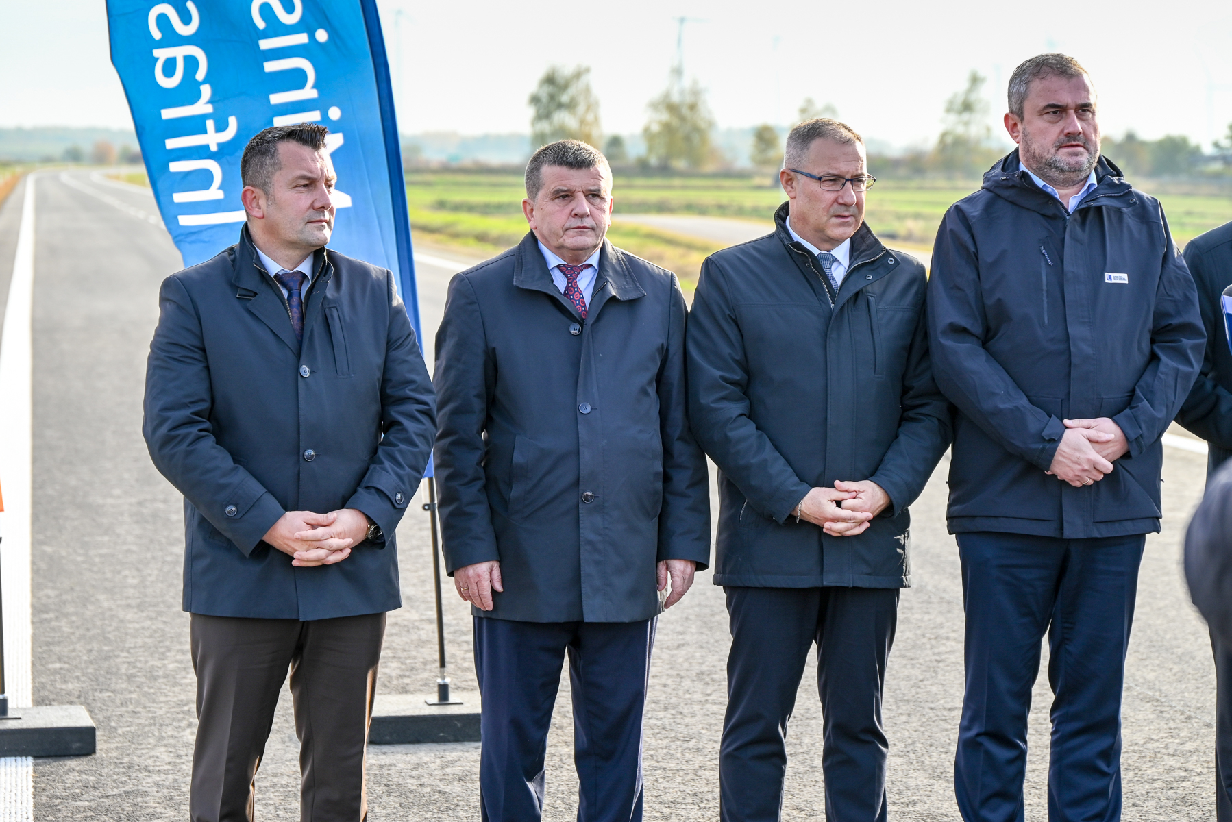 Czterech mężczyzn w ciemnych płaszczach i garniturach stoi na nowej drodze, w tle zielone pola i niebieska flaga z białym napisem.