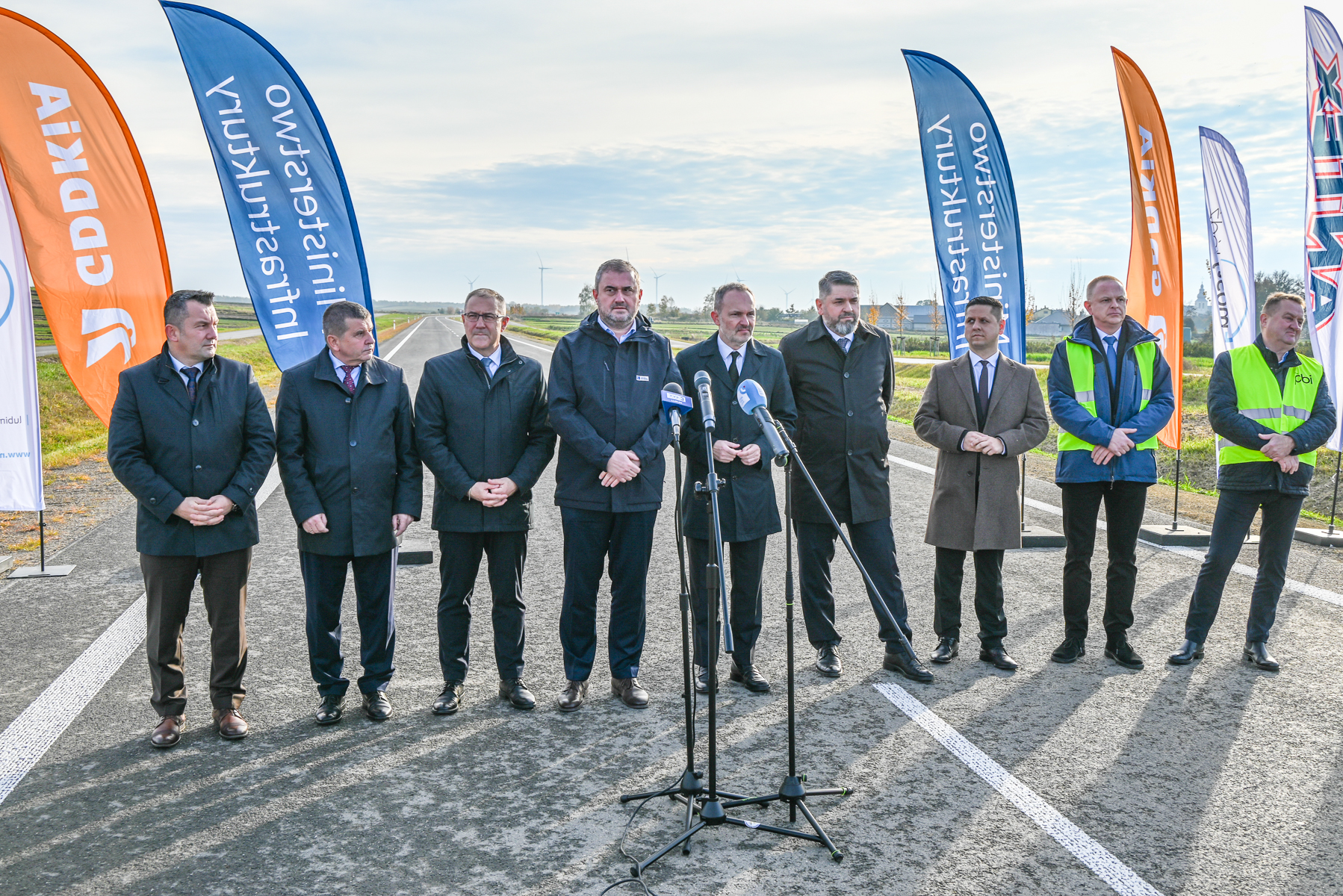 Dziesięciu mężczyzn w ciemnych płaszczach i garniturach stoi na drodze asfaltowej, za nimi kolorowe flagi reklamowe na tle nieba i zielonych pól.