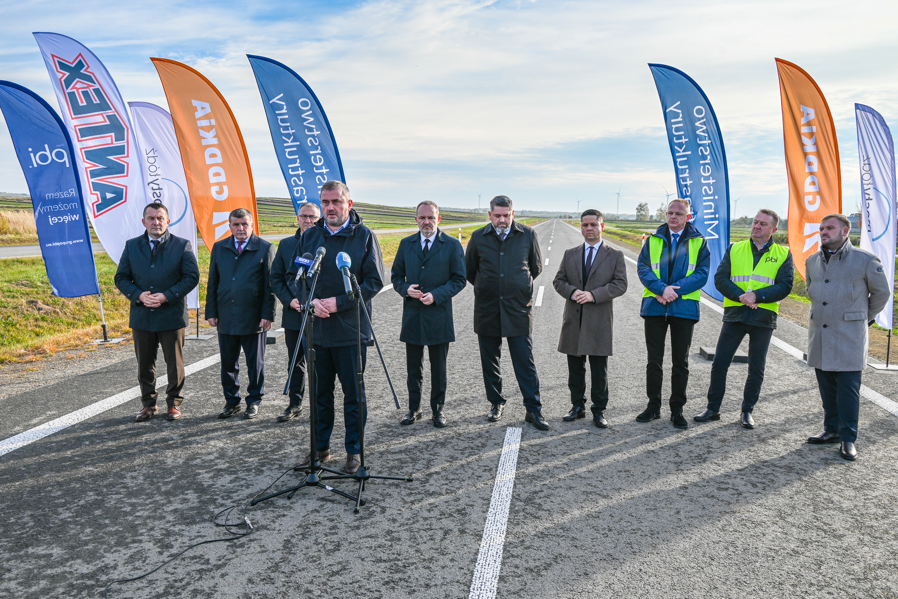 Dziesięciu mężczyzn w ciemnych płaszczach i dwóch w żółtych kamizelkach stoi na nowej drodze z mikrofonami, za nimi kolorowe flagi firmowe na tle nieba i pól.