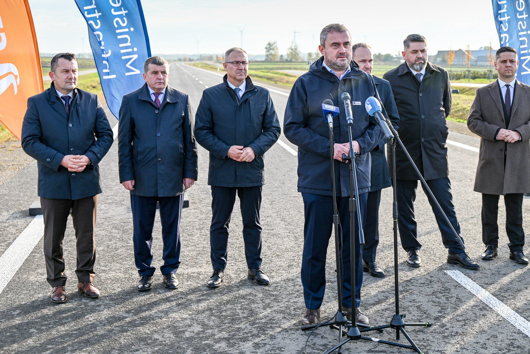 Siedmiu mężczyzn w ciemnych płaszczach stoi na nowej drodze, jeden mówi do mikrofonów, w tle flagi z napisami, zielone pola i wiatraki.