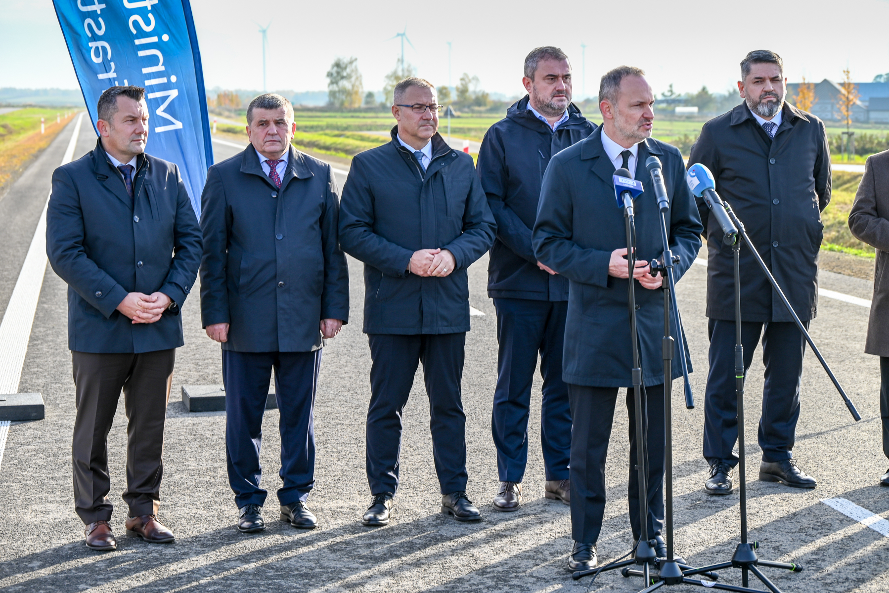 Sześciu mężczyzn w ciemnych płaszczach stoi na drodze, jeden mówi do mikrofonów, w tle zielone pola i wiatraki, obok niebieska flaga z białym napisem.