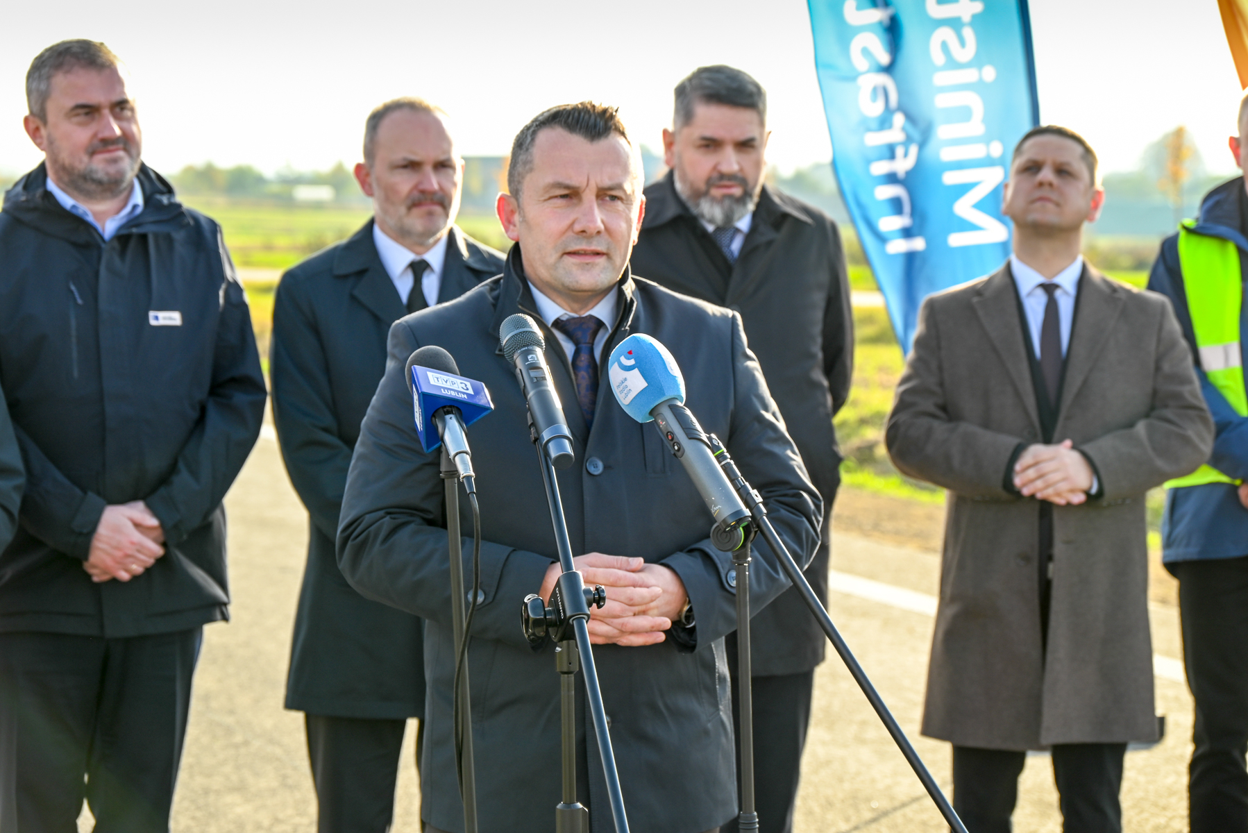 Pięciu mężczyzn w garniturach i płaszczach stoi na zewnątrz na drodze, jeden przemawia do mikrofonów, w tle niebieska flaga z napisem.