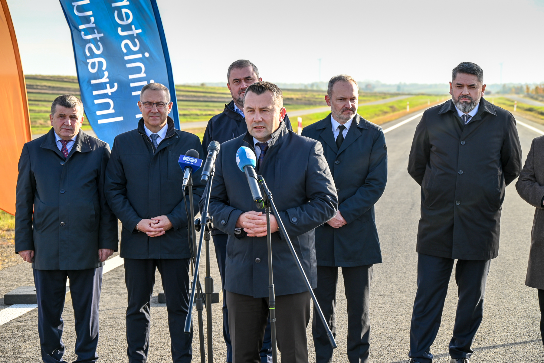 Sześciu mężczyzn w ciemnych płaszczach stoi na drodze, jeden przemawia do mikrofonów, w tle pola i niebo, z lewej strony widoczne niebieska i pomarańczowa flaga.