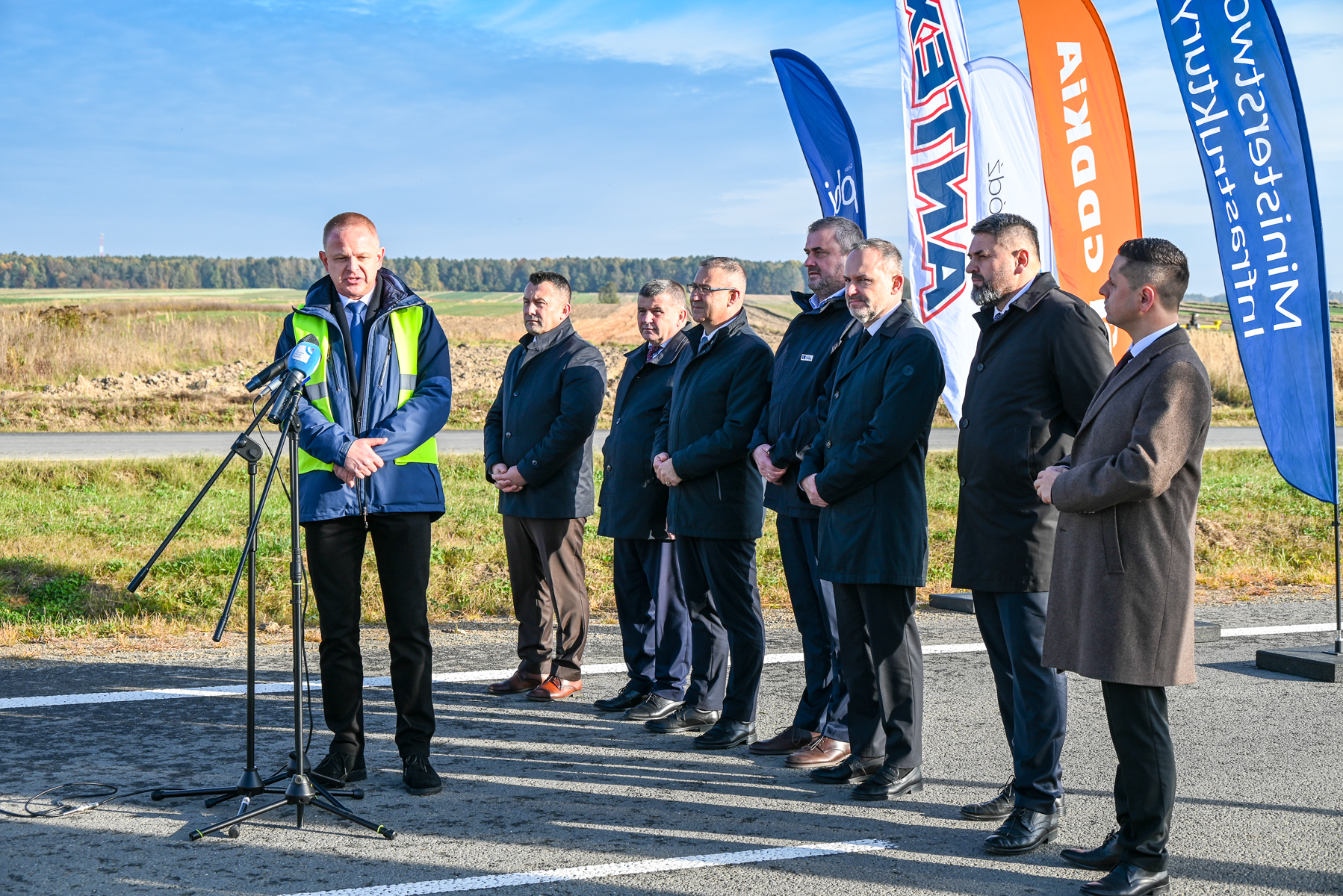 Grupa mężczyzn w ciemnych płaszczach i garniturach stoi na asfalcie, jeden z nich w kamizelce odblaskowej przemawia przy mikrofonach, w tle kolorowe flagi i zielona łąka.
