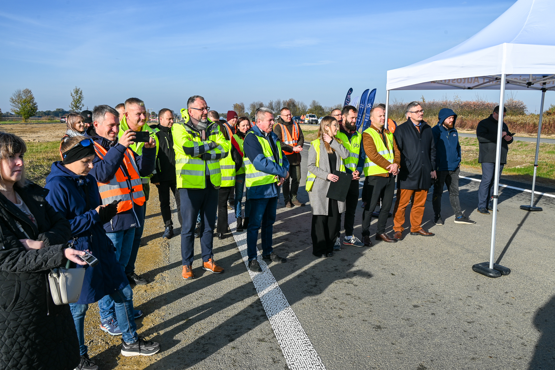Grupa ludzi w kamizelkach odblaskowych i kurtkach stojąca na drodze obok białego namiotu, na tle niebieskiego nieba i trawiastego terenu.