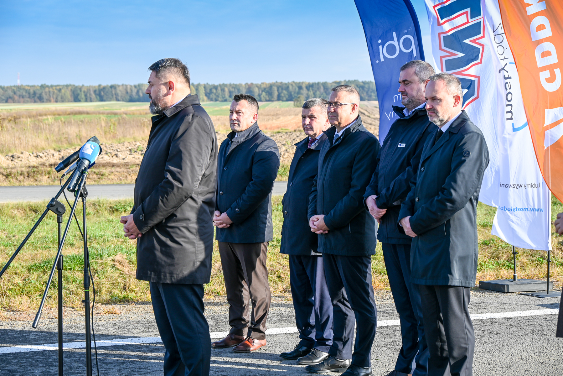 Sześciu mężczyzn w ciemnych płaszczach stoi na drodze asfaltowej, jeden przemawia przy mikrofonie, w tle zielone pola i trzy kolorowe flagi z napisami.