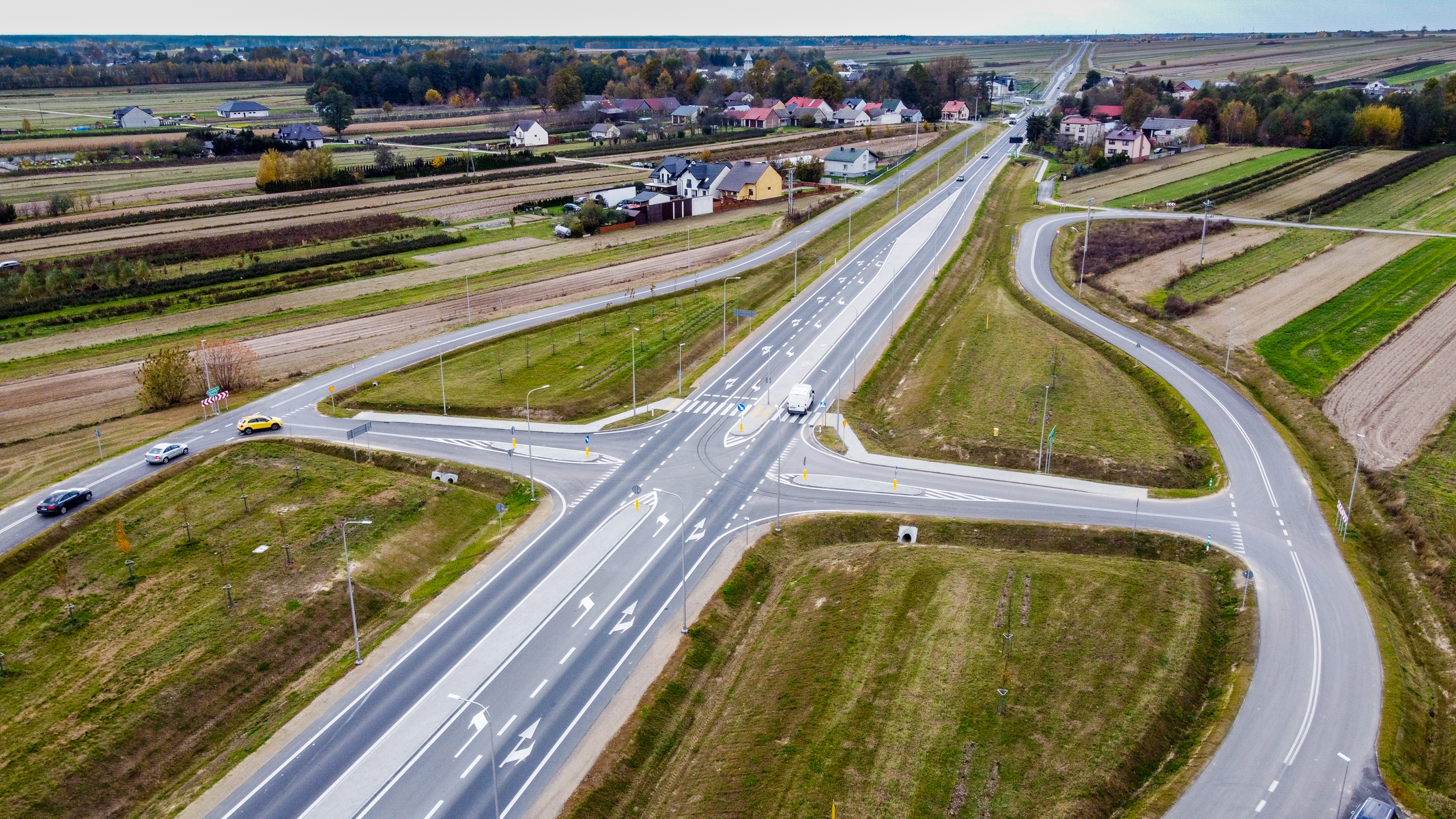 Skrzyżowanie wielopasmowe z oznaczeniami kierunków, białą ciężarówką i kilkoma samochodami, otoczone polami i domami na horyzoncie, jesienna zieleń i brązy.
