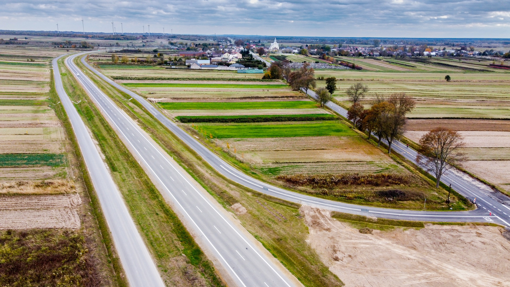 Widok z lotu ptaka na autostradę obok pól uprawnych o różnych odcieniach zieleni i brązu oraz małą wioskę w tle pod pochmurnym niebem.