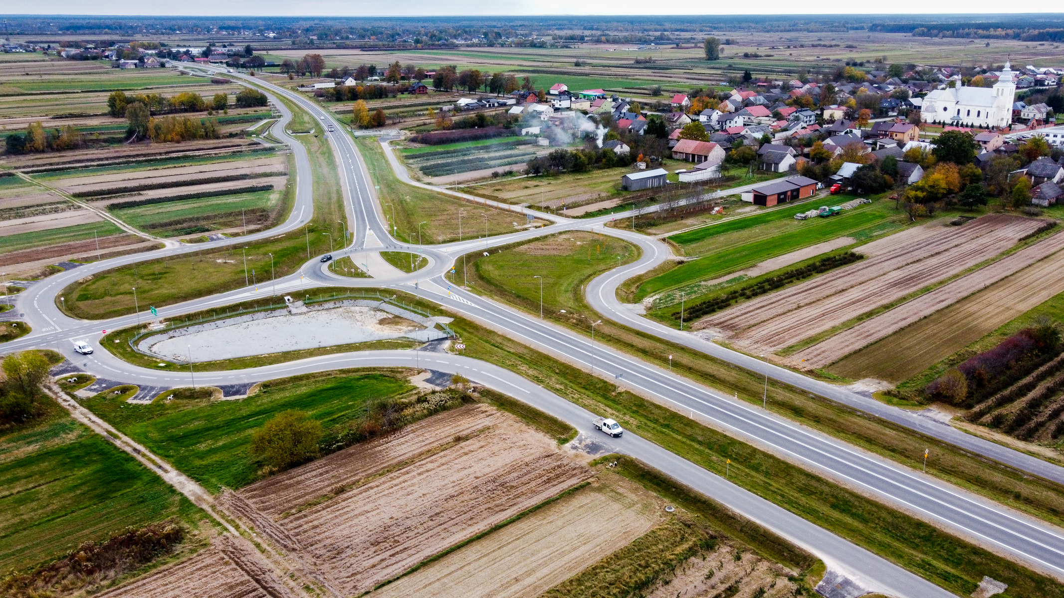 Skrzyżowanie z ronda i drogami otoczone zielonymi polami i uprawami, w tle zabudowa wiejska z domami i białym kościołem, jesienna pora.