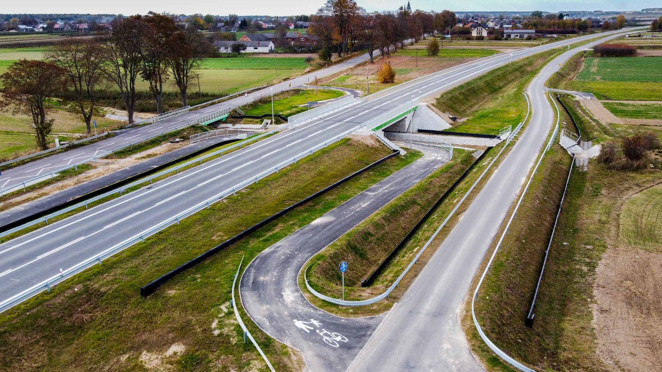 Skrzyżowanie dróg z mostem, asfaltowe jezdnie, ścieżka rowerowo-piesza z oznaczeniami, zielone pobocza, drzewa i zabudowania w tle.