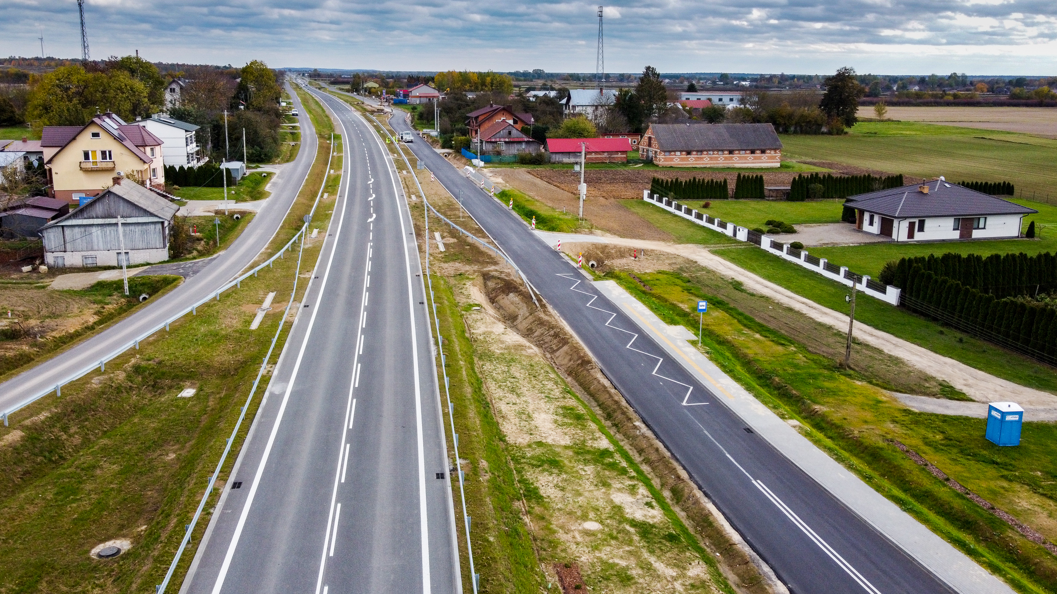 Widok z lotu ptaka na autostradę i boczną drogę, po bokach domy z ogrodami, zielona trawa i pola, pochmurne niebo.