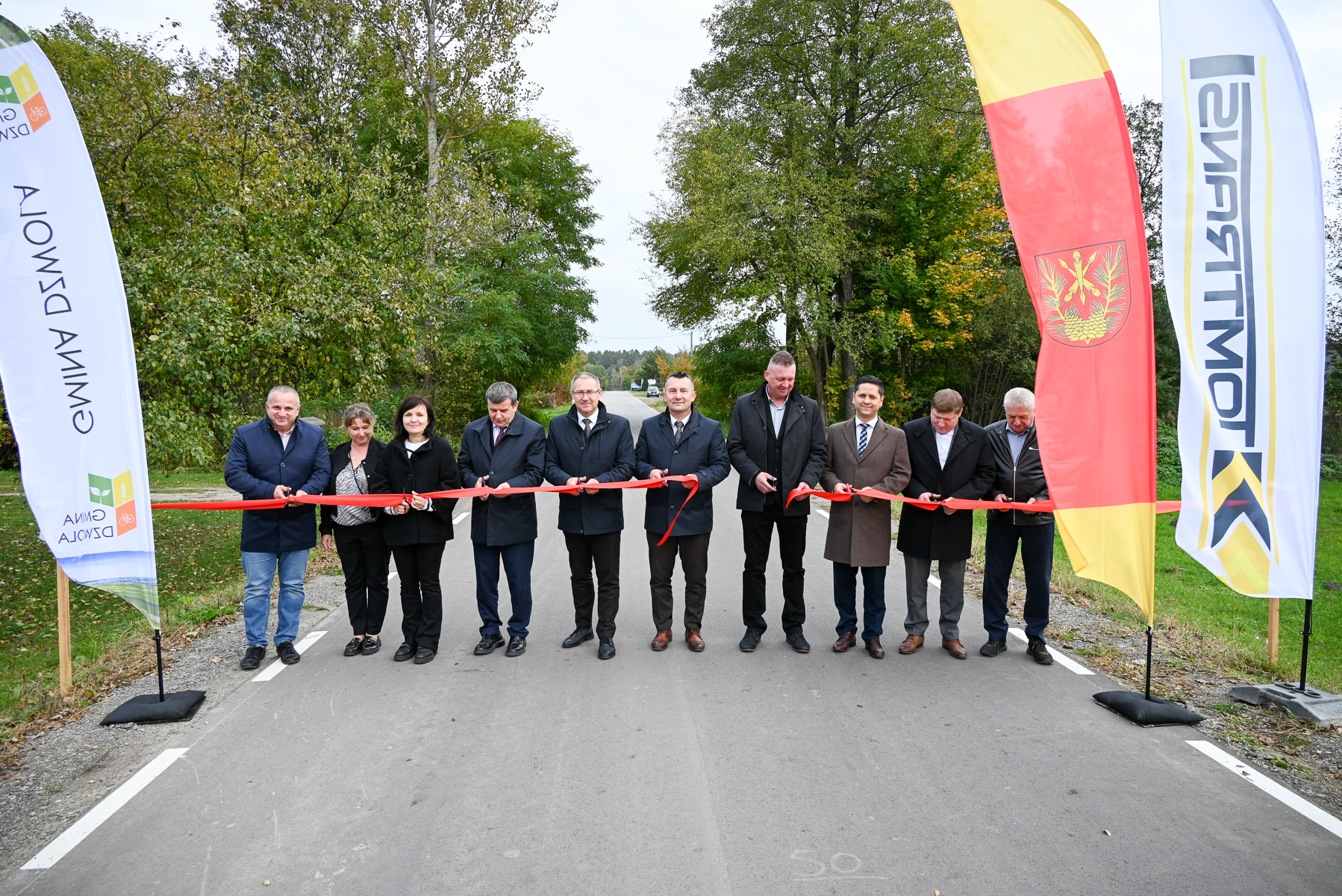 Dziesięć osób na asfaltowej drodze trzyma czerwone wstęgi, obok stoją trzy flagi, w tle drzewa i zarośla.