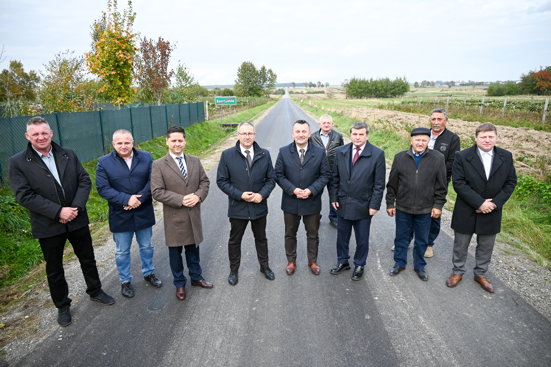 Dziesięciu mężczyzn w jesiennych płaszczach stoi na asfaltowej drodze na tle pól, drzew i nieba z chmurami. Po lewej zielony płot i tablica z nazwą miejscowości.