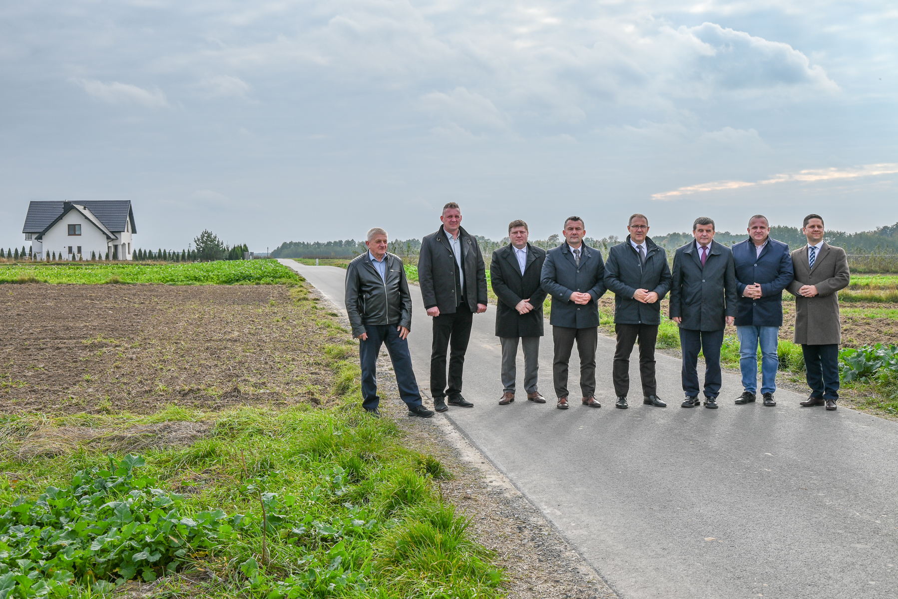 Siedmiu mężczyzn w ciemnych płaszczach stoi na asfaltowej drodze, po lewej stronie pole uprawne i biały dom z ciemnym dachem, pochmurne niebo.